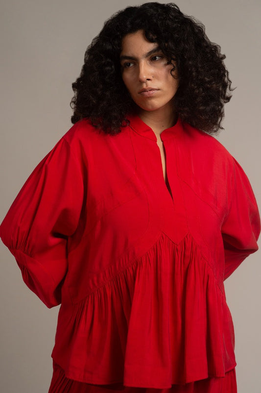 Una mujer talla XXL viste una blusa roja de manga larga con plisados hecha de cupro. La blusa es de la marca Yakampot, una firma de moda mexicana que fusiona tradición y modernidad . La blusa tiene un cuello redondo y un corte holgado que le da un toque de comodidad y estilo. La blusa es ideal para combinar con pantalones o faldas de colores neutros o contrastantes.